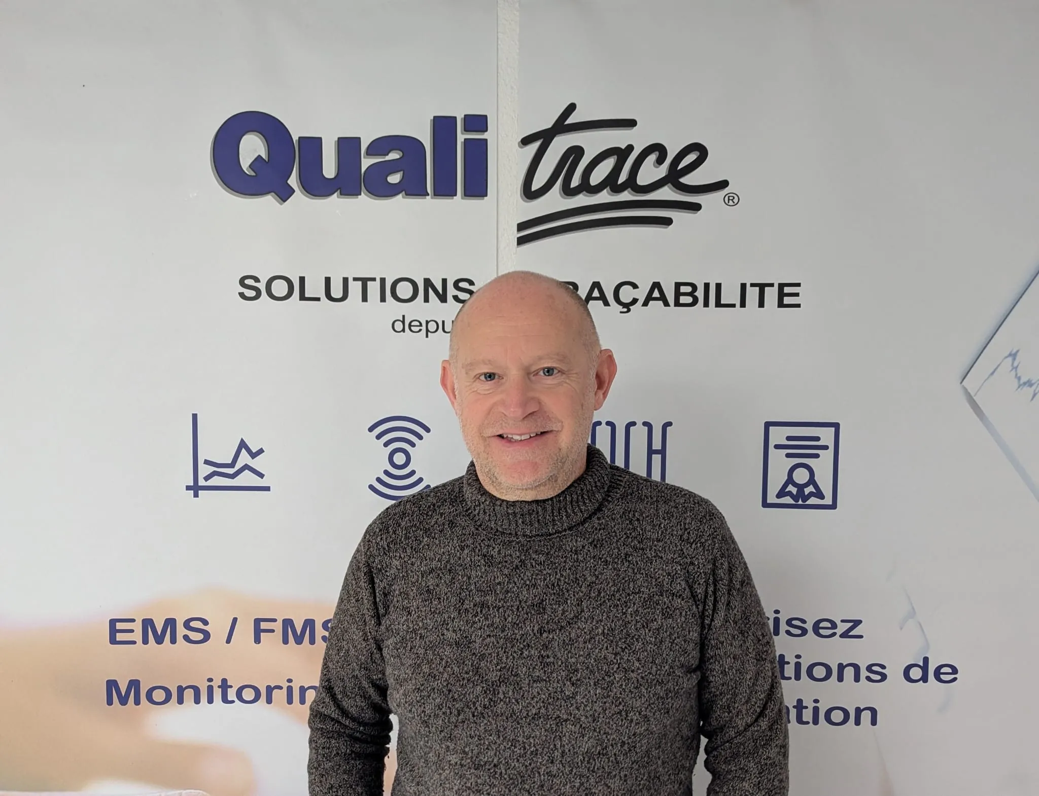 Portrait d’un membre de l’équipe devant une affiche Qualitrace, spécialisée dans les solutions de traçabilité et de monitoring environnemental.